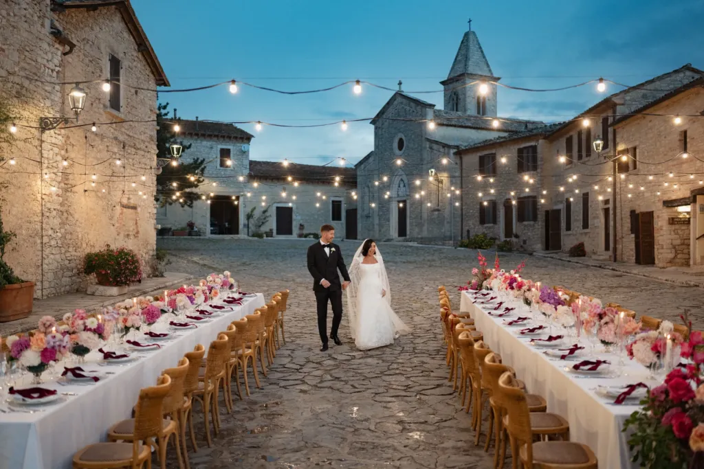 Bride and groom walking between long dinner tables in an Italian village square at dusk